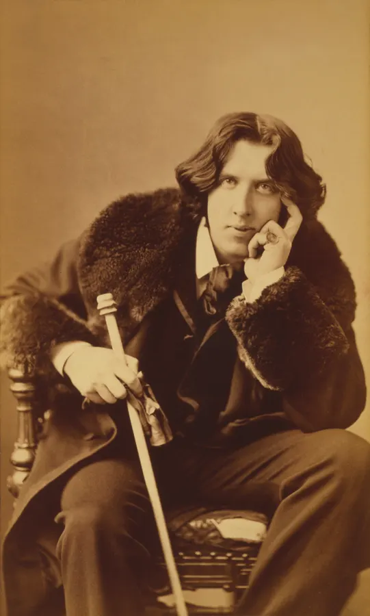 Oscar Wilde sitzt mit Mantel und Stock für ein Porträt von Napoleon Sarony